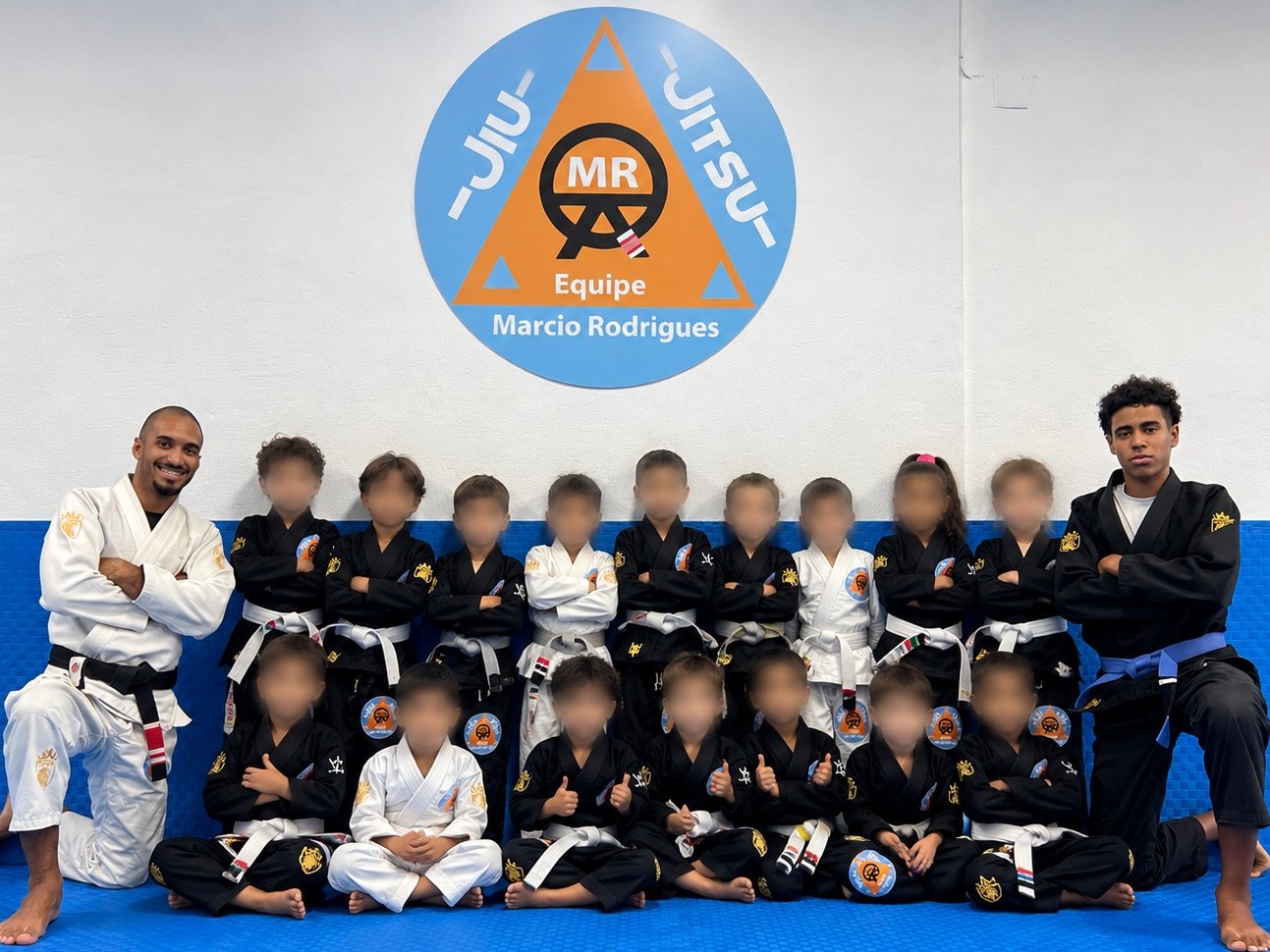Grupo de crianças dos 3 aos 6 anos, em kimonos com faixas brancas, ajoelhado no tatame azul com os rostos desfocados
